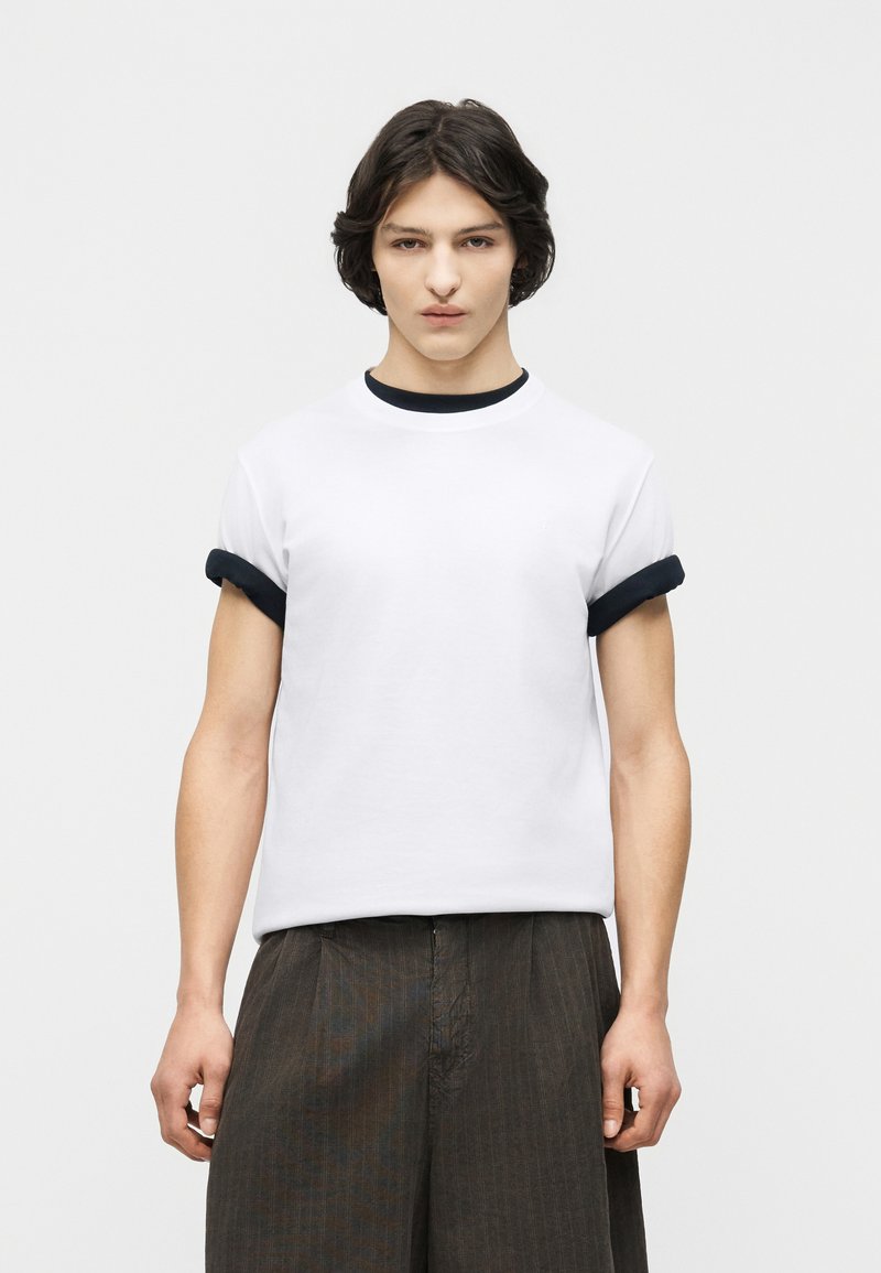 Νεαρό άτομο με σκούρα μαλλιά που φοράει λευκό μπλουζάκι με μαύρη λεπτομέρεια και φαρδιά σκοτεινά παντελόνια με ρίγες, στέκεται μπροστά σε απλό φόντο.