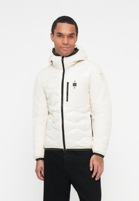 Veste isolée blanche avec un design matelassé, capuche ajustée, fermetures éclair noires et une poche poitrine. Caractéristiques des poignets élastiques et un ourlet côtelé.