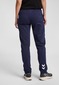 Marinblå träningsbyxor med elastisk midja, smalnande ben och vit chevronranddetalj nära vristerna. Bärs med svarta sneakers.