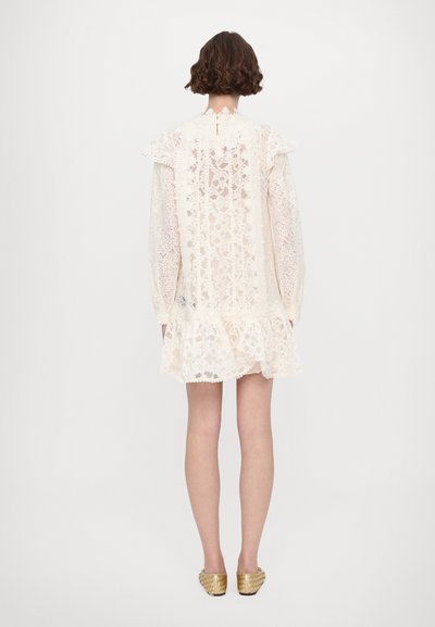 Spitzenkleid in Creme mit langen Ärmeln und Rüschenausschnitt. Besteht aus filigranen Blumenmustern und einem gestuften Saum, kombiniert mit goldenen Slippers.
