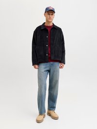 Black corduroy jacket with front buttons, red knitted sweater underneath, light blue jeans, tan footwear, and a blue cap with logo.