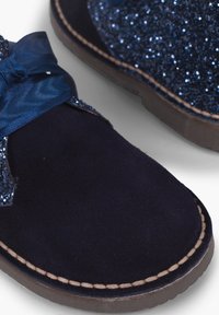 Botines de ante azul marino con un acento brillante azul, una cinta de satén y una suela cosida en contraste para un detalle adicional.