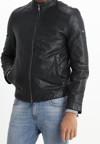 Man in een zwart leren jack met een voorrits en een blauwe spijkerbroek, staand met één hand in zijn zak en de andere ontspannen.