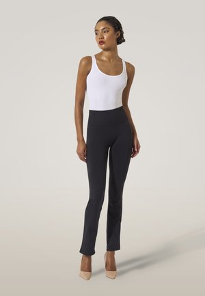 Vrouw met kort haar in een witte top, zwarte high-waisted broek en beige hakken, staande met haar handen langs haar zijde tegen een effen achtergrond.