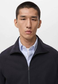 Black zip-up fleece jacket with a high collar over a light blue collared shirt. Smooth texture with a modern cut and clean lines.