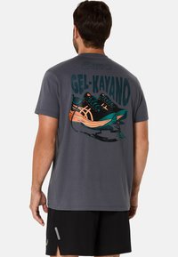 Grå T-shirt med korte ærmer, der har et farverigt grafik af løbesko og teksten "GEL-KAYANO" på ryggen. Lavet af et blødt stof.
