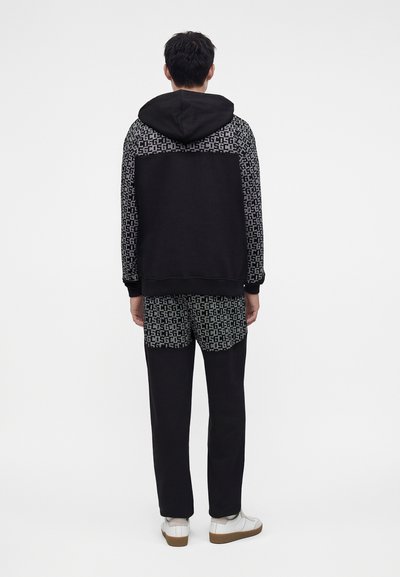 Homme debout de dos portant un sweat à capuche noir et gris à motifs et un pantalon assorti avec des baskets blanches sur fond blanc.
