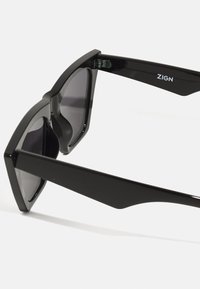 Ochelari de soare negri cu un design geometric, având lentile întunecate, rame groase și o formă ascuțită la tâmple. Logo-ul brandului pe braț.