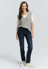 Chaleco de punto acanalado gris sobre una camisa blanca, combinado con jeans azul oscuro y zapatillas beige. El modelo está de pie en un fondo neutro.