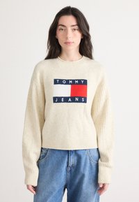 Jasno beżowy sweter z prążkowanymi rękawami i dołem, z niebieskim, białym i czerwonym prostokątnym logo z napisem "TOMMY JEANS."