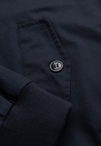 Gros plan sur un tissu foncé avec une poche cousue et un bouton noir étiqueté "OMBRE", à côté d'un poignet côtelé d'une manche longue.