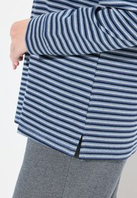 Maglietta a righe con linee diagonali blu e bianche, con uno spacco laterale all'orlo. Abbinata a pantaloni grigi a trama.