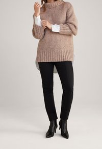 Maglione rosa chiaro in maglia grossa con colletto alto, orlo a coste e spalle scese, indossato sopra una camicia bianca; abbinato a pantaloni neri attillati e stivali.
