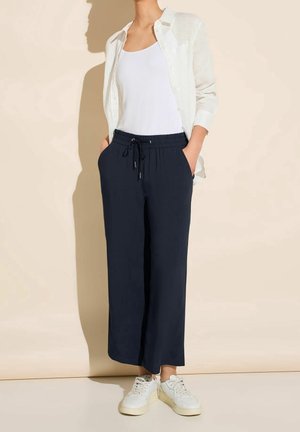 Pantaloni elasticizzati blu navy con tasche laterali, abbinati a una canotta bianca e una camicia leggera a bottoni con maniche lunghe.