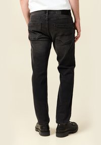 Hombre vestido con jeans negros y zapatos de cuero negros, de espaldas a la cámara contra un fondo beige.