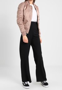 Ljusrosa bomberjacka med en slät textur, vit t-shirt under, svarta byxor med vid ben, och svarta sneakers med vita sulor.