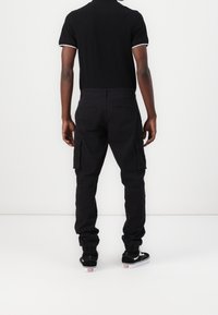 Schwarze Cargohosen mit mehreren Taschen, an den Knöcheln eng anliegend, kombiniert mit einem schwarzen Poloshirt mit weißen Akzenten und schwarzen Sneakern.