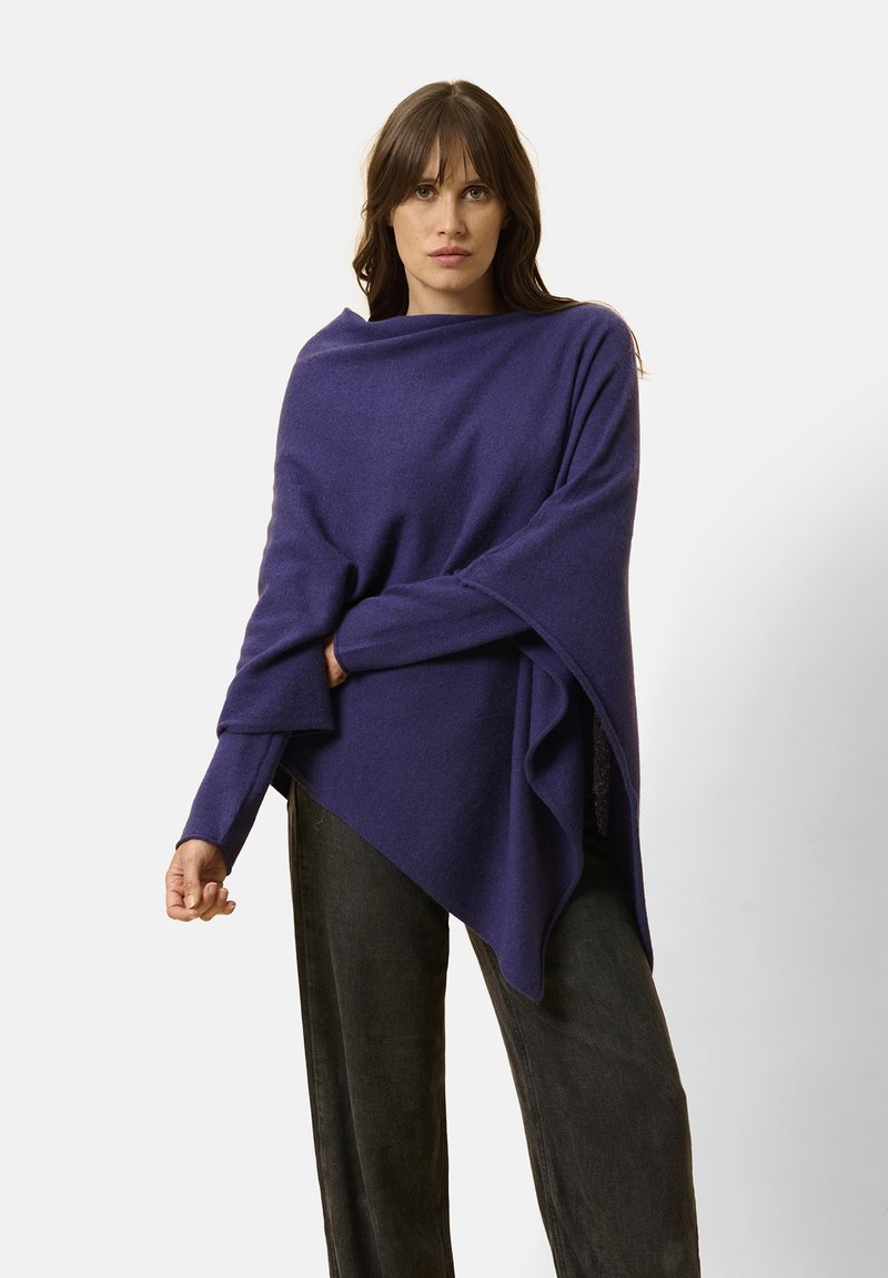 Fioletowy wełniany poncho o luźnym kroju, asymetrycznym wzorze, długich rękawach oraz teksturowanej tkaninie spływającej na przodzie. Połączony z ciemnymi spodniami.