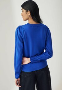 Pull en maille bleu avec des manches bouffantes, des poignets et un ourlet côtelés, texture lisse, associé à un jean foncé. Vue de l'arrière.