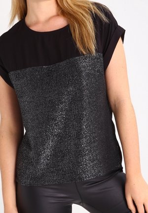 Femme portant un haut noir à manches courtes avec un panneau texturé et brillant sur le devant et un pantalon en cuir noir.