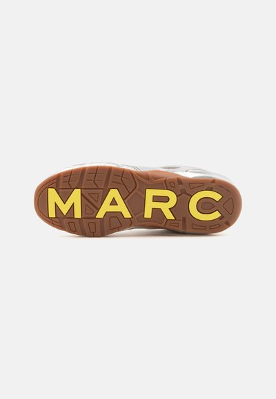 Semelle d'une basket blanche avec une bande de roulement marron et de grandes lettres jaunes épelant "MARC" sur le dessous.