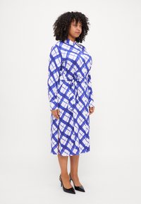 Abito camicia a scacchi blu e bianchi con maniche lunghe, colletto, vita legata e spacchi laterali, abbinato a decolté nere a punta.