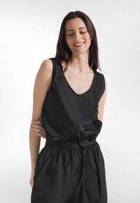 Schwarzes ärmelloses Oberteil mit tiefem V-Ausschnitt, aus glattem Stoff, weit geschnitten. Kombiniert mit passenden schwarzen Hosen mit elastischem Bund.