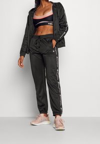 Černý sportovní outfit zahrnující zipovou bundu, sportovní podprsenku s barevnými bloky a zúžené tepláky s bočními kapsami a logovými akcenty.