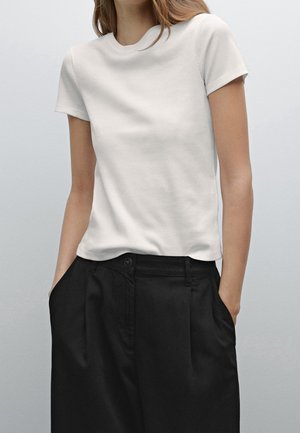 Kvinde iført en enkel hvid kortærmet ribbet T-shirt, der er stoppet ned i sorte højtaljede bukser med hænderne i lommerne.