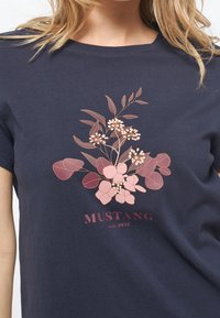 Marinblus med rosa blommönster och texten "MUSTANG est. 1932" på bröstet, bärs av en person med blont hår.