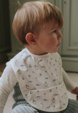 Bambino piccolo con capelli castano chiaro che indossa un bavaglino bianco decorato con piccoli leopardi e palme, seduto all’interno e guarda verso destra.