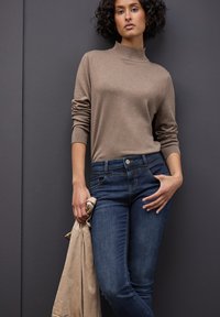 Beige högkragad tröja gjord av mjukt tyg, tillsammans med mörkblå denimjeans. Ljusbrun jacka hängande över armen.