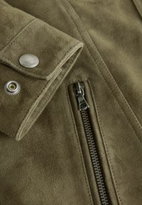 Artimos planas alyvuogių žalios spalvos zomšinės striukės, kurioje matyti sulankstytas rankogalis su sidabrine spaudės saga ir vertikali juoda užtrauktuku ant minkštos tekstūruotos medžiagos.