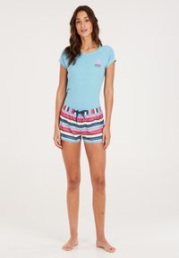Světle modré tričko s krátkým rukávem a malou růžovou a modrou grafikou, spárované s barevně pruhovanými plavkami s elastickým pasem.