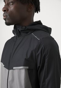 Veste de sport noire et grise avec capuche, dotée d'une fermeture éclair, d'accents réfléchissants et de la marque 'Ellesse' sur l'épaule. Texture lisse.