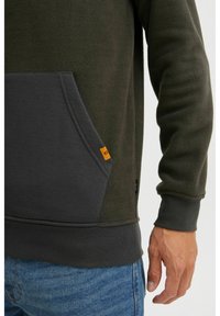 Sudadera de forro polar verde oscuro con un bolsillo gris, que presenta una etiqueta de acento naranja. La textura es suave y esponjosa, con puños y dobladillo acanalados.