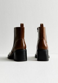 Bottines en cuir marron avec un talon épais noir, dotées d'une fermeture éclair à l'arrière et d'une petite languette, texture lisse et design simple.