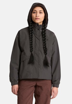 Frau trägt eine dunkel Kapuzenjacke und braune Hosen mit zwei langen geflochtenen Zöpfen und steht vor einem hellen Hintergrund.