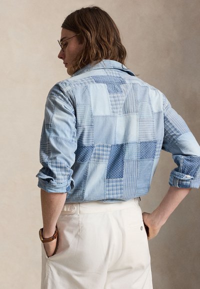 Personne portant des lunettes et une chemise en jean patchwork retroussée aux tons bleus, avec un pantalon blanc, debout les mains sur les hanches, de profil.