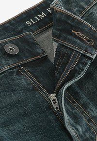 Slim fit traperice od tamnoplave tkanine, s vidljivim šavovima, metalnim patentnim zatvaračem i teksturiranim pojasom s oznakom.