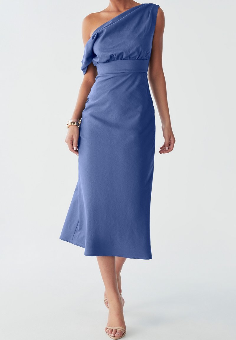 Femme portant une robe midi bleue sans manches, asymétrique, avec des talons ouverts beige et des bracelets dorés, debout devant un fond uni.