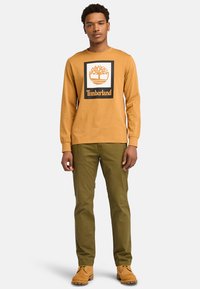 Orange long-sleeve shirt featuring a black-bordered graphic logo. Olive green pants and tan boots complete the outfit.