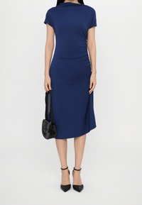 Mörkblå figurnära klänning med korta ärmar och rynkade detaljer, knälång, i samklang med en liten svart handväska och spetsiga svarta klackskor.