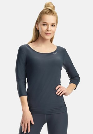 Blondínka s účesom s vrchným uzlom, oblečená v priliehavom tmavosivom tričku s trojštvrťovým rukávom a legínach, pózujúca s jednou rukou na bedre.