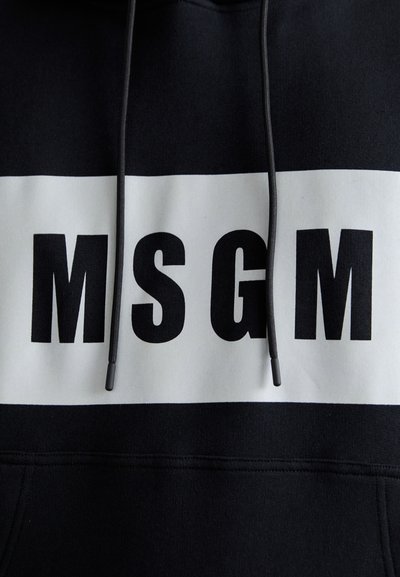 Schwarzer Hoodie mit einem auffälligen weißen horizontalen Streifen, auf dem die Buchstaben "MSGM" in großen schwarzen Lettern stehen. Der Hoodie hat schwarze Kordeln mit Plastikspitzen.