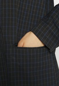 Černá kostkovaná tkanina s modrými a hnědými pruhy, s rukou v boční kapse, ukazující na precizní design a čisté linie.