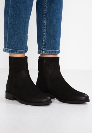 Czarne zamszowe botki za kostkę z bocznym zamkiem, noszone z niebieskimi podwiniętymi jeansami na białej podłodze i tle.