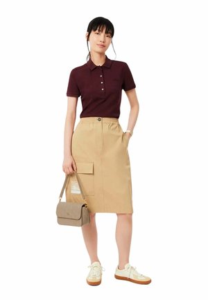 Polo shirt bordeaux avec un col, jupe beige jusqu'aux genoux avec une taille élastique et une poche latérale, baskets crème et un sac à main léger.