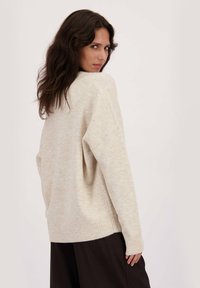 Vrouw met lang donker haar, gekleed in een losse beige trui en zwarte broek, iets gedraaid met een neutrale uitdrukking tegen een effen achtergrond.