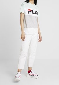 Camiseta corta verde claro y gris con el logo "FILA" en negro y rojo, combinada con jeans blancos y zapatillas blancas con detalles en rojo.